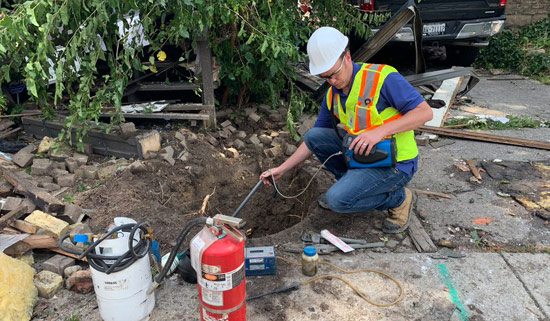 A technician wearing a white hard hat and a high-visibility vest crouches beside a small, excavated hole in the ground, using a handheld device with a probe to inspect an underground line. A red fire extinguisher and other equipment are visible on the disturbed ground nearby.