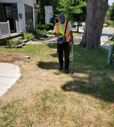 A male Hetek technician wearing a reflective safety vest and cap walks across a residential lawn, operating a handheld gas leak detection device with a long probe during a pipeline inspection.